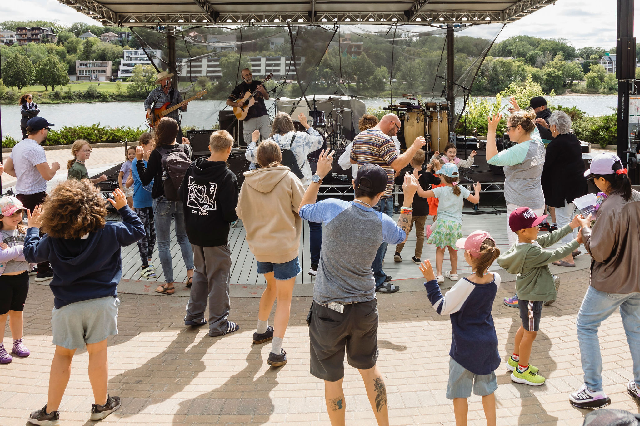 A group of children and adults dance together in front of a live outdoor band performance on a riverside stage. The scene is lively and joyful, with people of all ages enjoying the music on a sunny day.