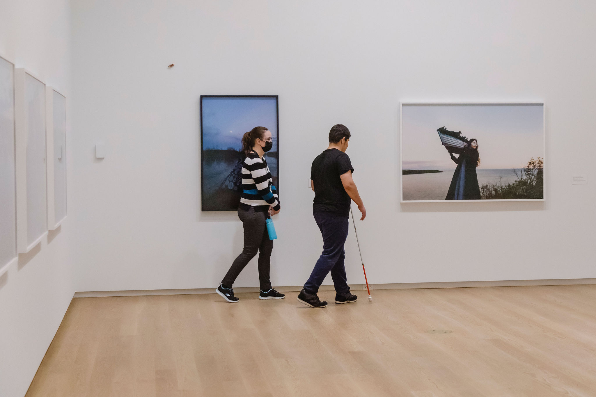 Two people walk through an art gallery; one is using a white cane for navigation and the other wears a mask and carries a water bottle. They pass framed photographs on the wall, including an image of a woman holding a structure by the sea. The space has light wooden floors and white walls.
