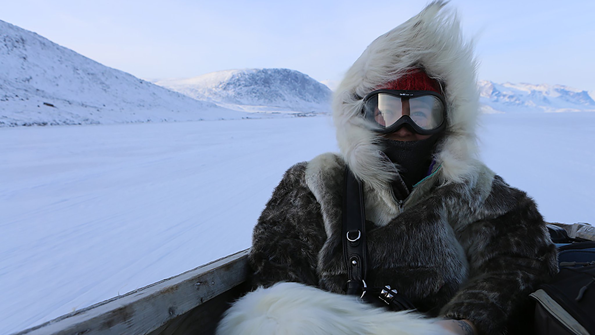 A person bundled in a thick fur parka with a large hood sits in a wooden sled on a snowy landscape. They wear ski goggles, a red headband, and a black face covering. Snow-covered mountains rise in the distance under a pale blue sky.