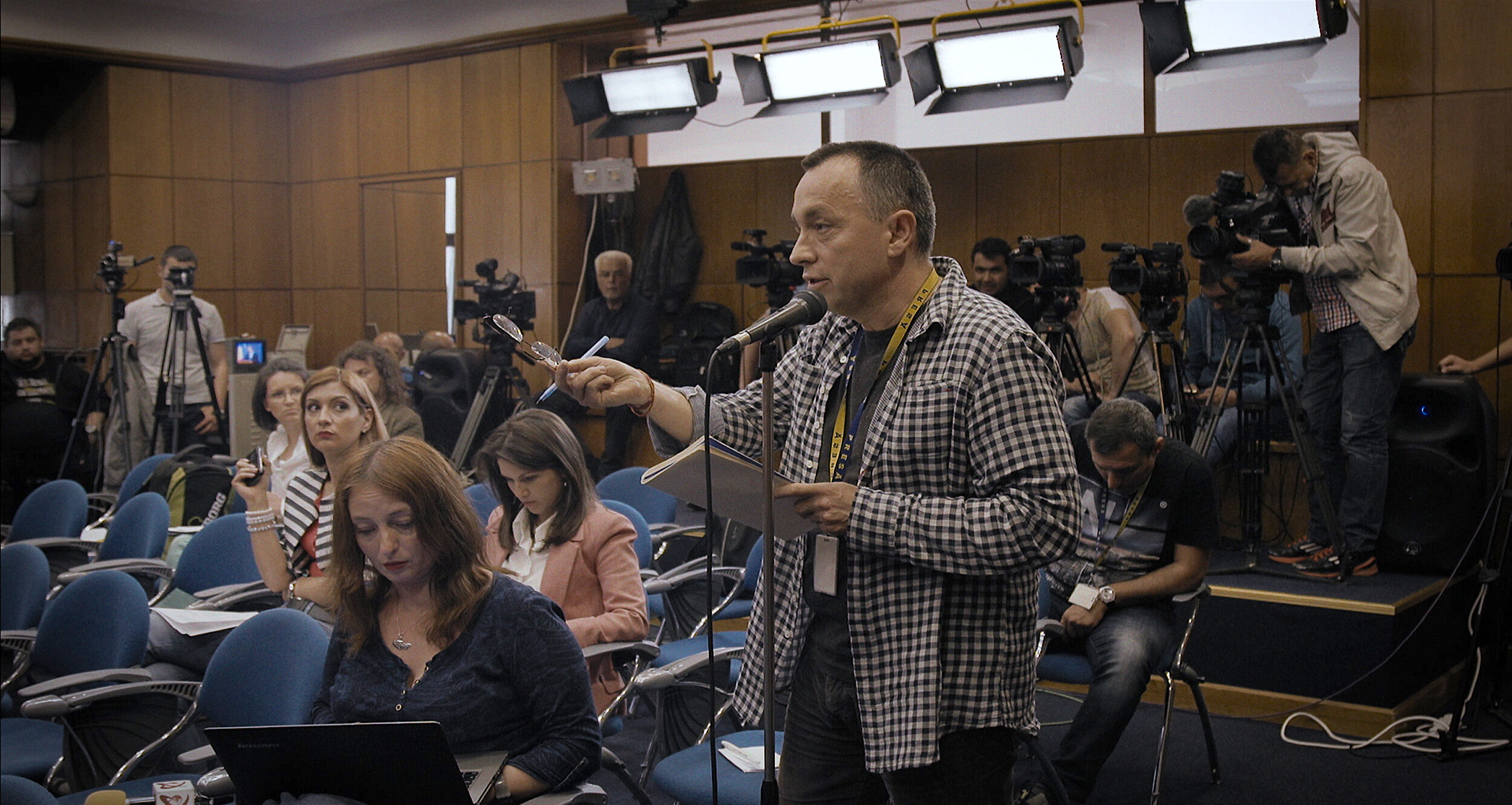 A man stands holding glasses and a notebook while speaking into a microphone during a press conference in a wood-paneled room. Behind him, journalists sit in rows of blue chairs with laptops and notepads, while camera operators record the event under bright studio lights.