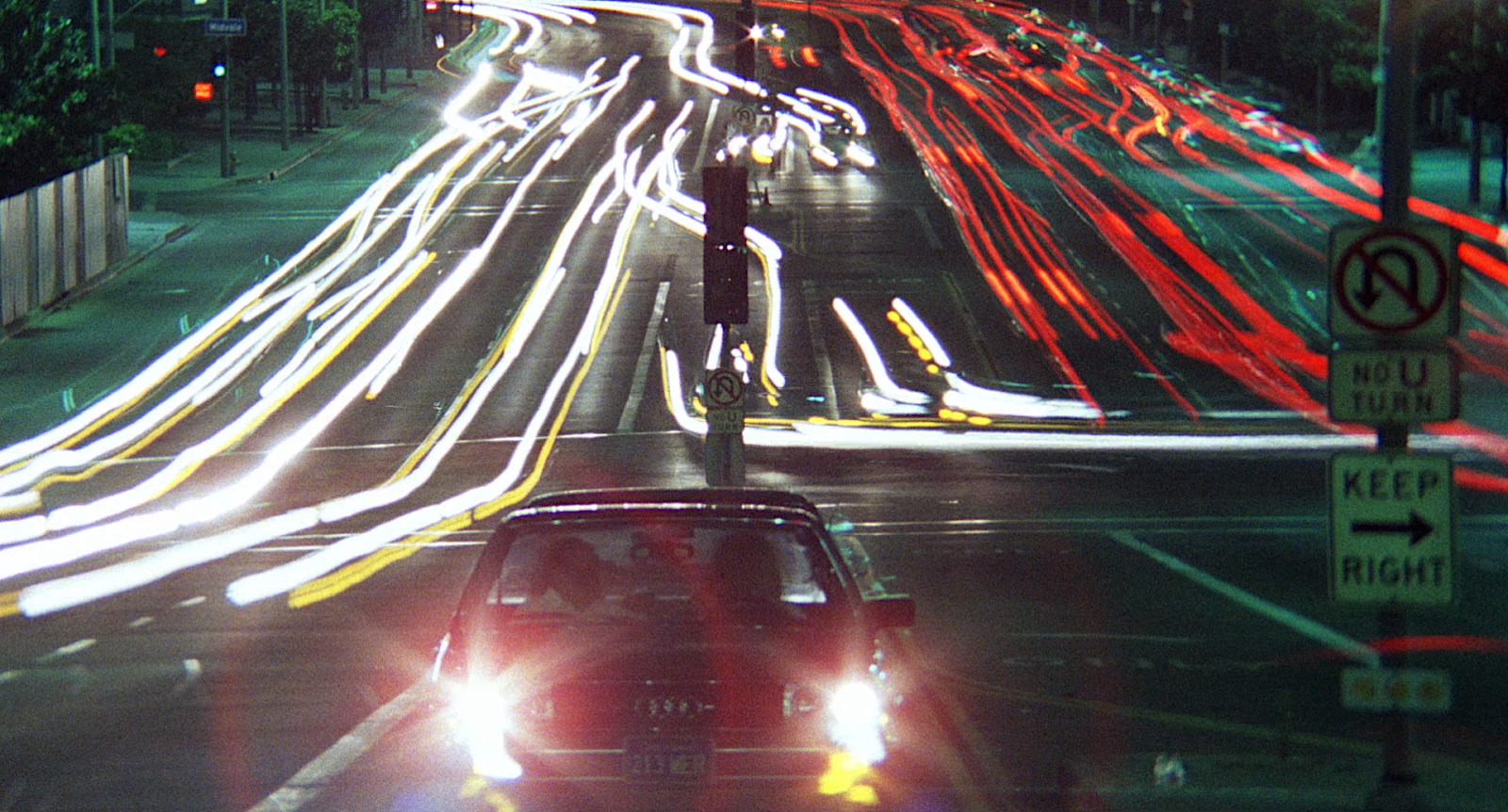 A night-time long exposure photograph of a city intersection with streams of red and white light trails created by moving vehicles. In the foreground, a stationary car with headlights on faces the camera. Street signs on the right side read "NO U TURN" and "KEEP RIGHT." The surrounding buildings and trees are dimly lit.