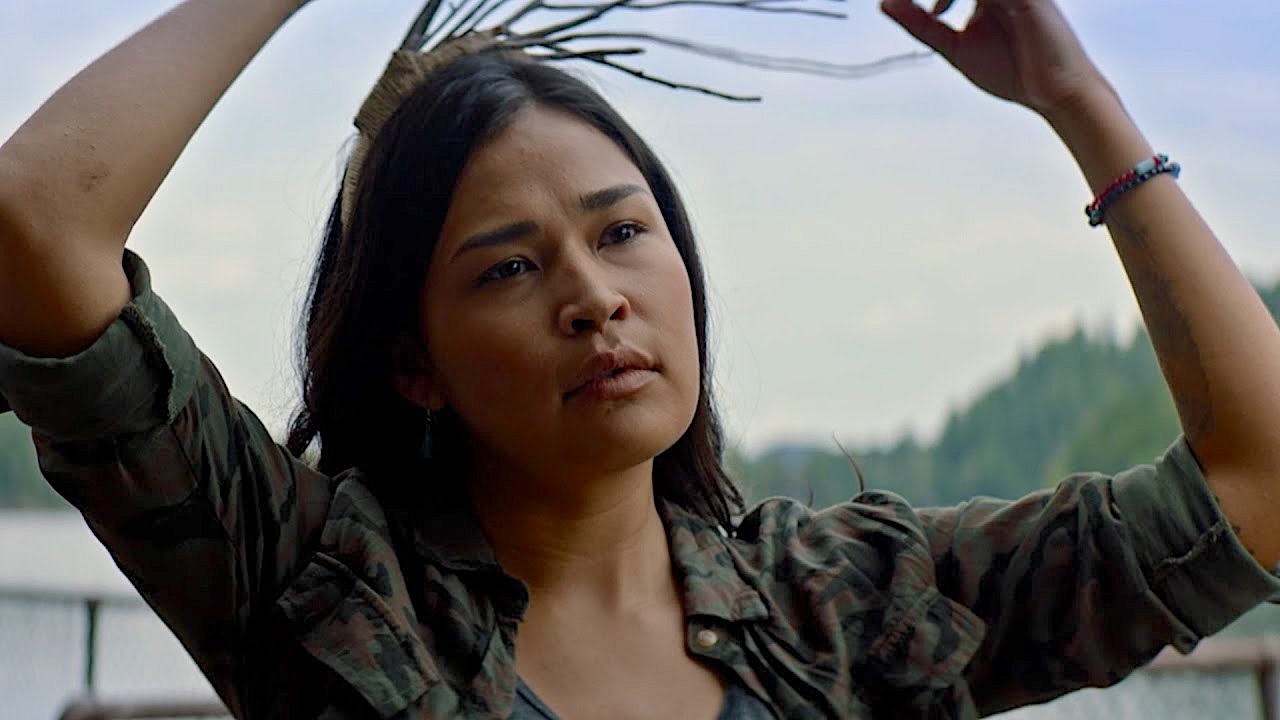 A woman with long dark hair and a thoughtful expression raises her arms to adjust a small woven headdress with sticks. She wears a camouflage jacket over a grey shirt and a beaded bracelet. In the background are trees and a calm lake under a cloudy sky.