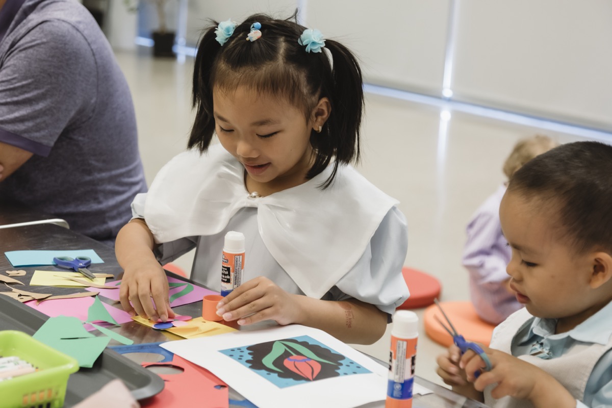 Two young children sit at a table engaged in a colourful paper art activity. One child smiles while arranging cut-out shapes with a glue stick, while the other concentrates on using small scissors. Bright craft materials are scattered across the table.
