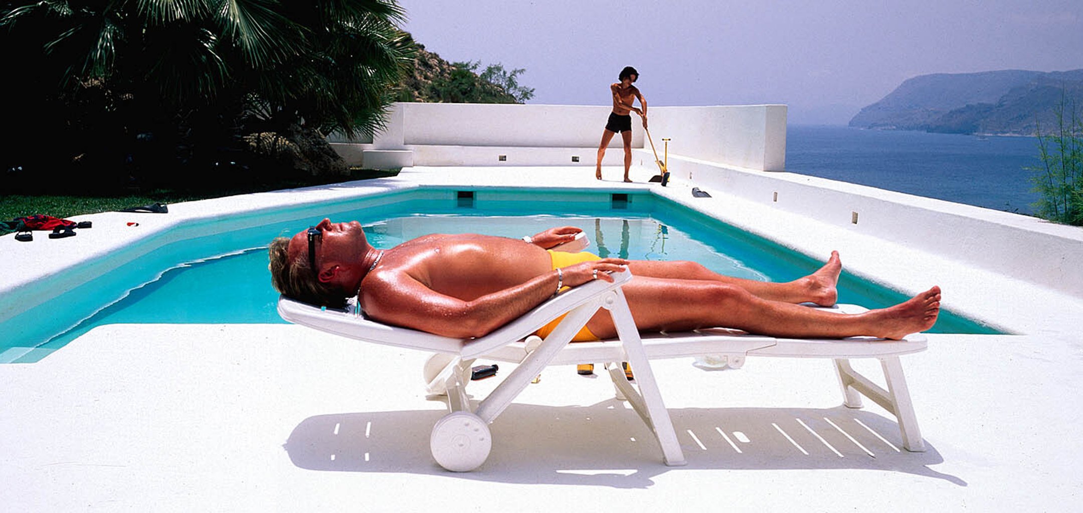A shirtless man wearing yellow swim trunks and sunglasses lounges on a white pool chair beside a turquoise swimming pool. In the background, a person in black shorts sweeps the white pool deck. The setting overlooks an ocean bay with distant hills under a hazy sky. Palm trees and lush greenery border the modern white terrace.