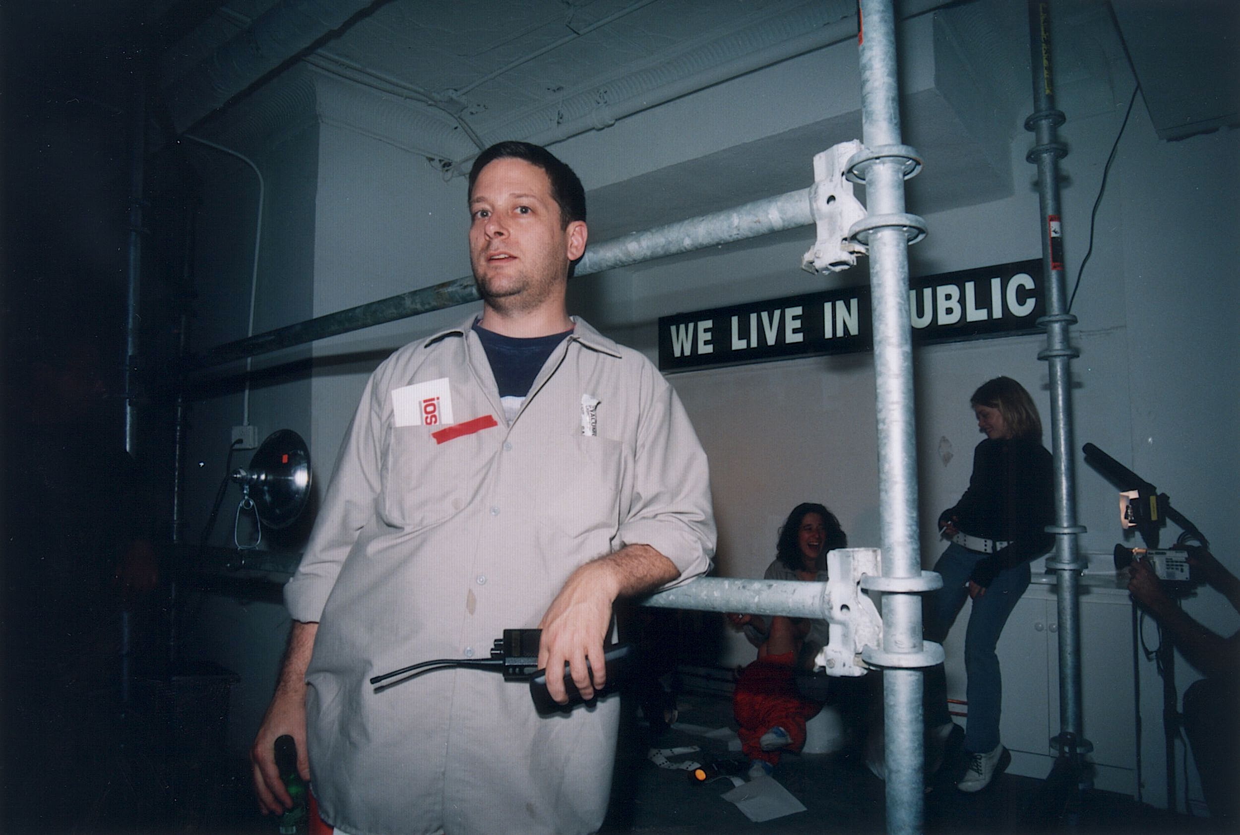A man in a light grey jumpsuit with a red name tag stands leaning against a metal scaffold in an industrial-looking room. He holds a small electronic device with wires. Behind him, a black sign with white letters reads “WE LIVE IN PUBLIC.” In the background, two women are interacting—one is sitting and laughing while the other stands nearby. A camera operator films the scene from the right.