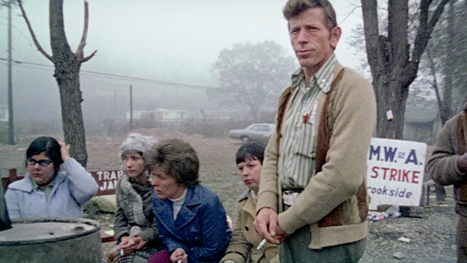 A man stands in front of a group of seated women outdoors on a foggy day. He wears a beige cardigan and striped shirt, holding a cigarette. Behind them is a sign reading “U.M.W.A. ON STRIKE Brookside,” indicating a miners’ strike. The women sit near a barrel and appear engaged in conversation.