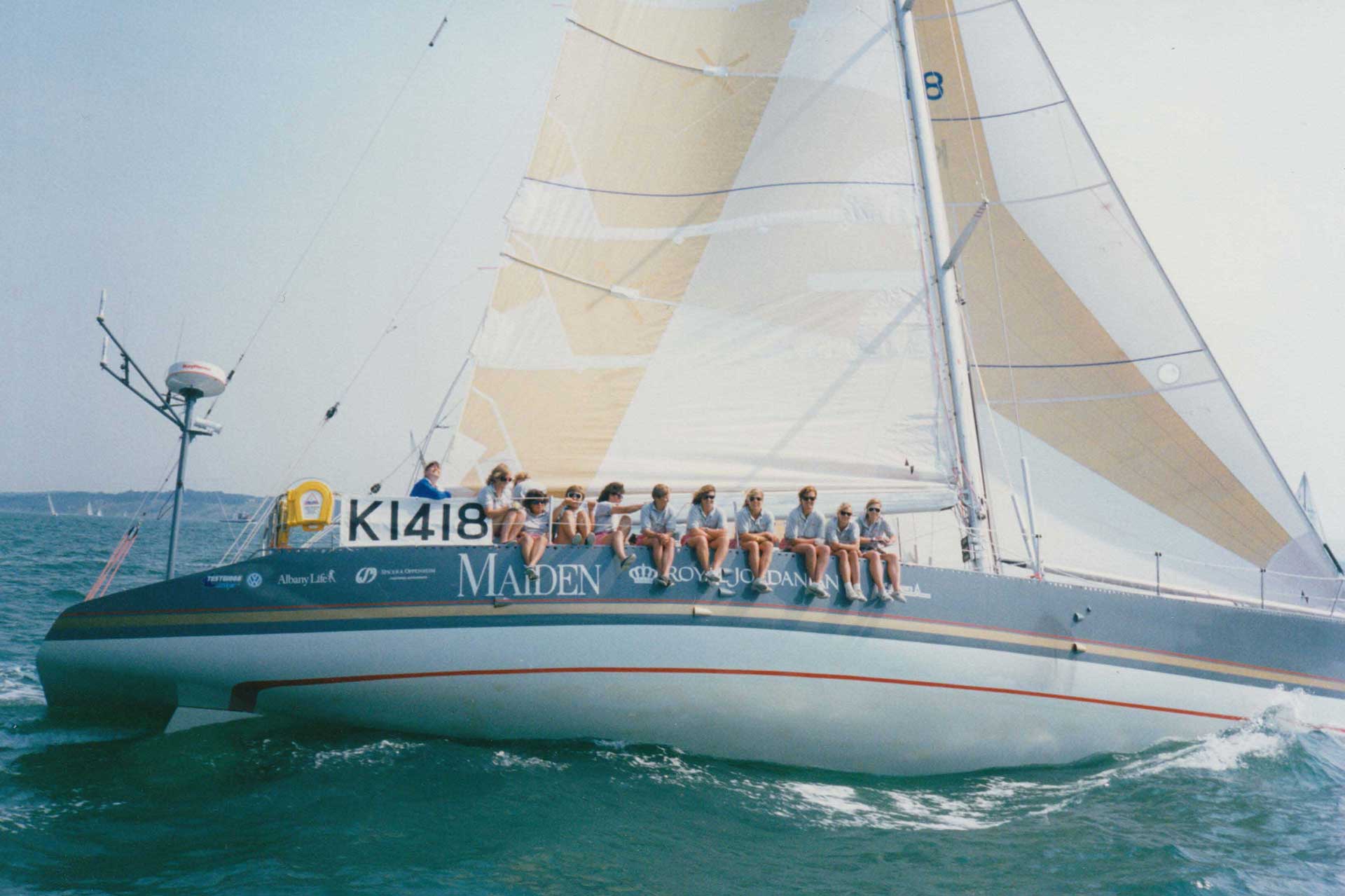 A sailboat named Maiden glides across the water under full sail. A team of women sits along the edge of the boat, legs dangling over the side, wearing sunglasses and sailing gear. The boat has a large sail with cream and white sections and displays sponsor logos. The sky and sea are clear and blue.