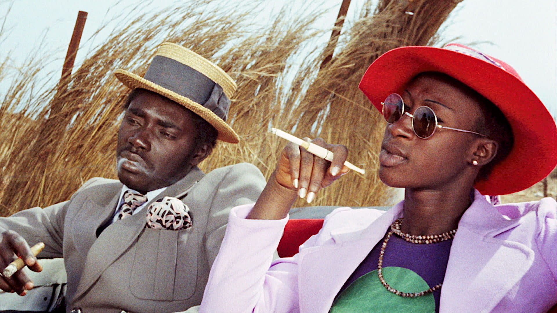 A stylish man and woman sit in an open car, wearing bright, fashionable outfits. The man in a light suit and straw hat smokes a cigar, while the woman in a red wide-brim hat, sunglasses, and lavender blazer holds a cigarette holder. Tall grass fills the background.