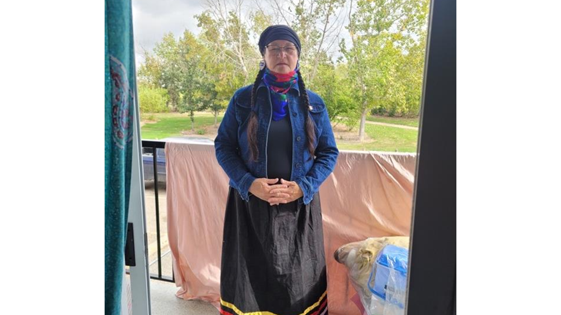 A person stands on a balcony outdoors, wearing a dark headscarf, colorful scarf, denim jacket, and long black skirt with red, yellow, and purple stripes at the hem. Their hands are clasped in front, and they are looking forward with eyes closed. Trees and a grassy park are visible in the background.