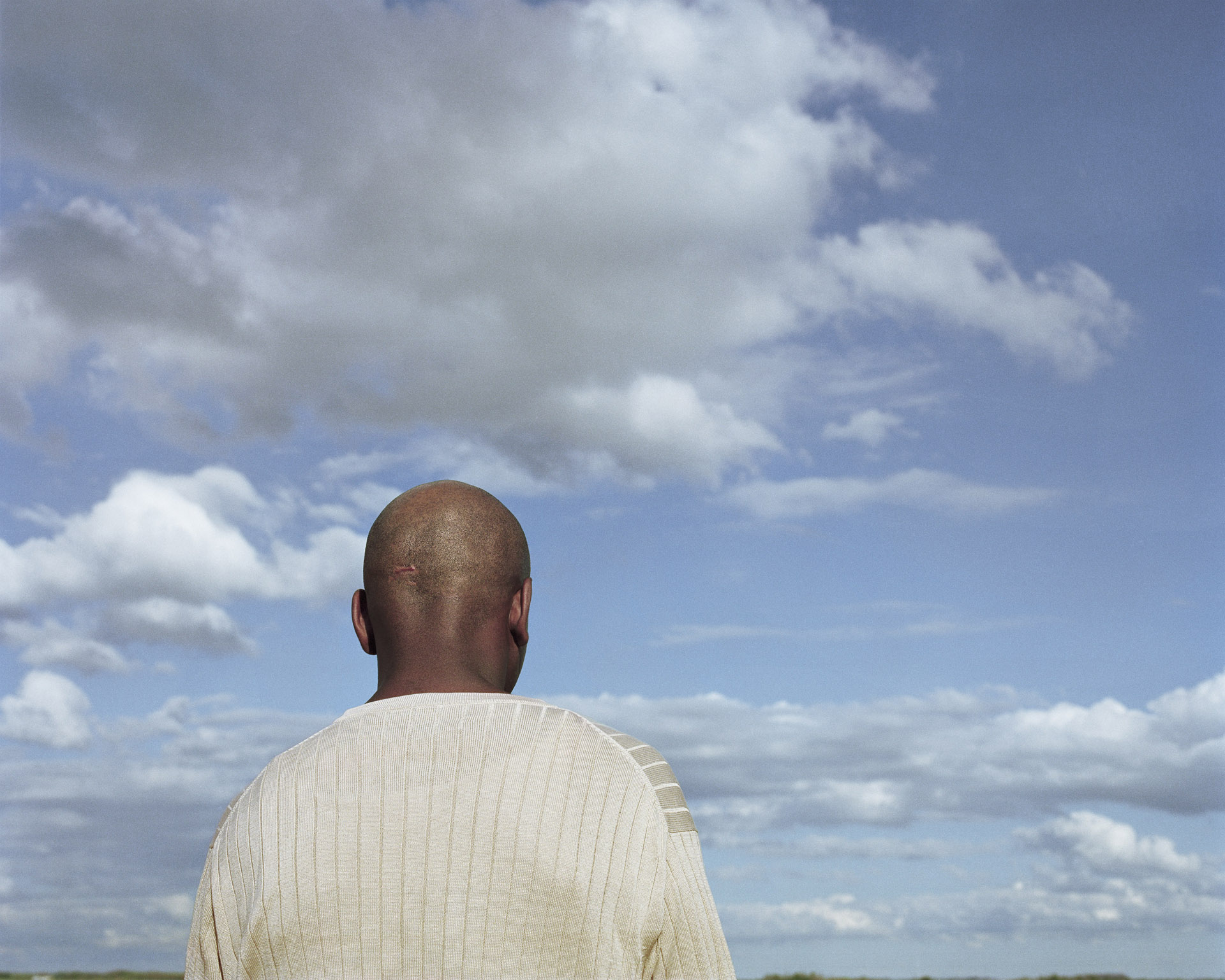 A man with a shaved head, seen from behind, stands beneath a vast, partly cloudy sky. He wears a cream-colored ribbed sweater, and a small scar is visible on the back of his head. The landscape below is mostly obscured, emphasizing the expansive sky above.