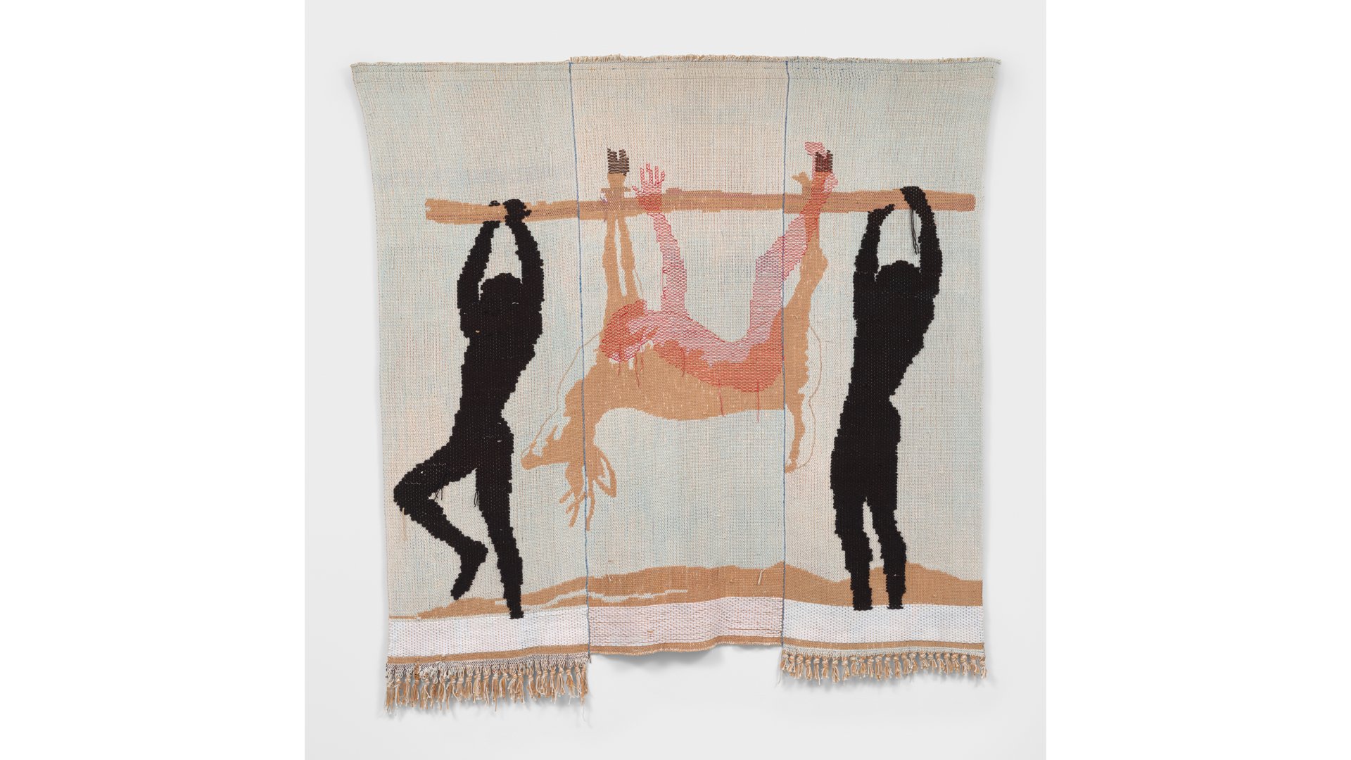 A woven textile depicting two black silhouetted figures carrying a deer upside down on a wooden pole. The deer is rendered in beige and pink tones, its legs tied to the pole, with its head and antlers hanging downward. The background is a pale blue with a sandy ground and fringed edges at the bottom.