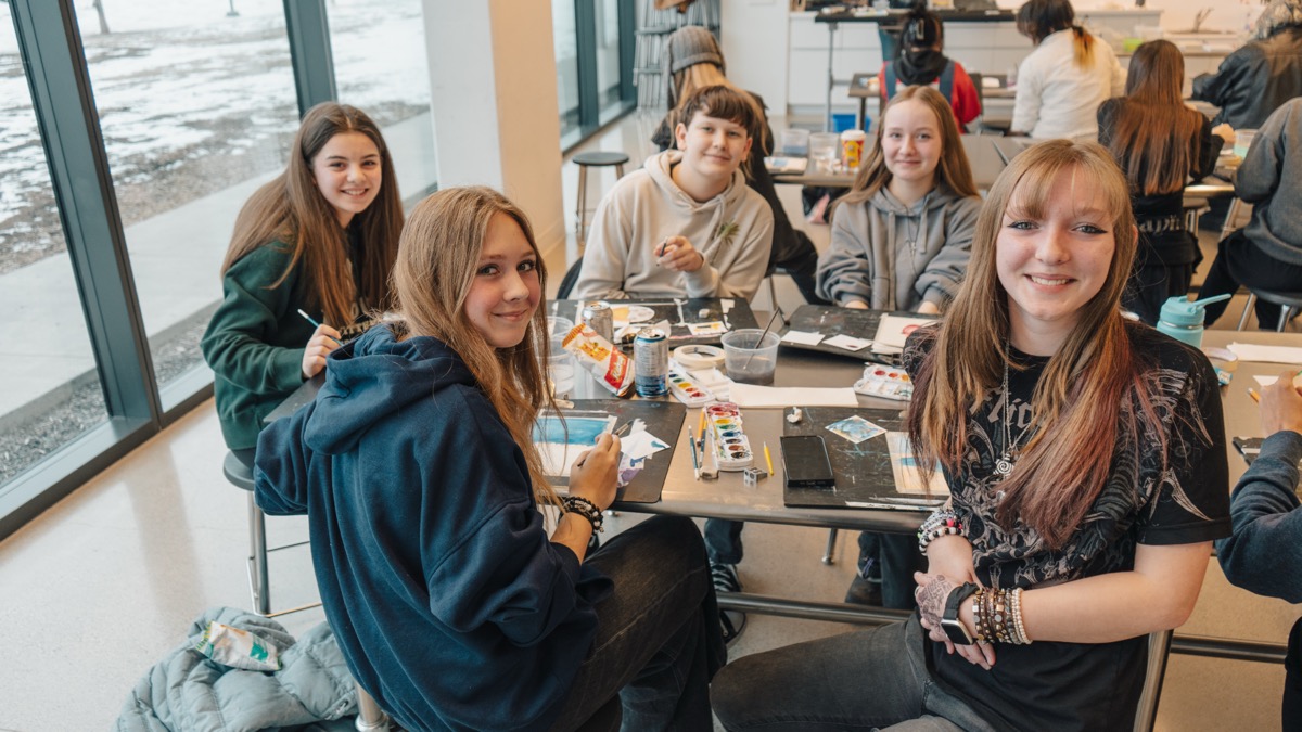 A group of smiling teenagers sit around a table in a bright art studio, painting with watercolours and enjoying snacks. Art supplies, paint palettes, and brushes are spread across the table. Large windows in the background reveal a snowy outdoor scene.