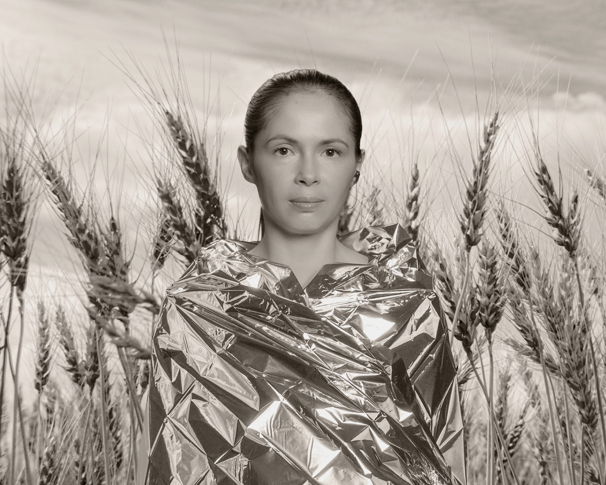 A person stands in a wheat field wearing a reflective metallic blanket that shimmers in the light. The portrait is in black and white, with tall wheat stalks surrounding the person under a softly clouded sky. The overall mood feels calm and contemplative, blending natural and futuristic elements.