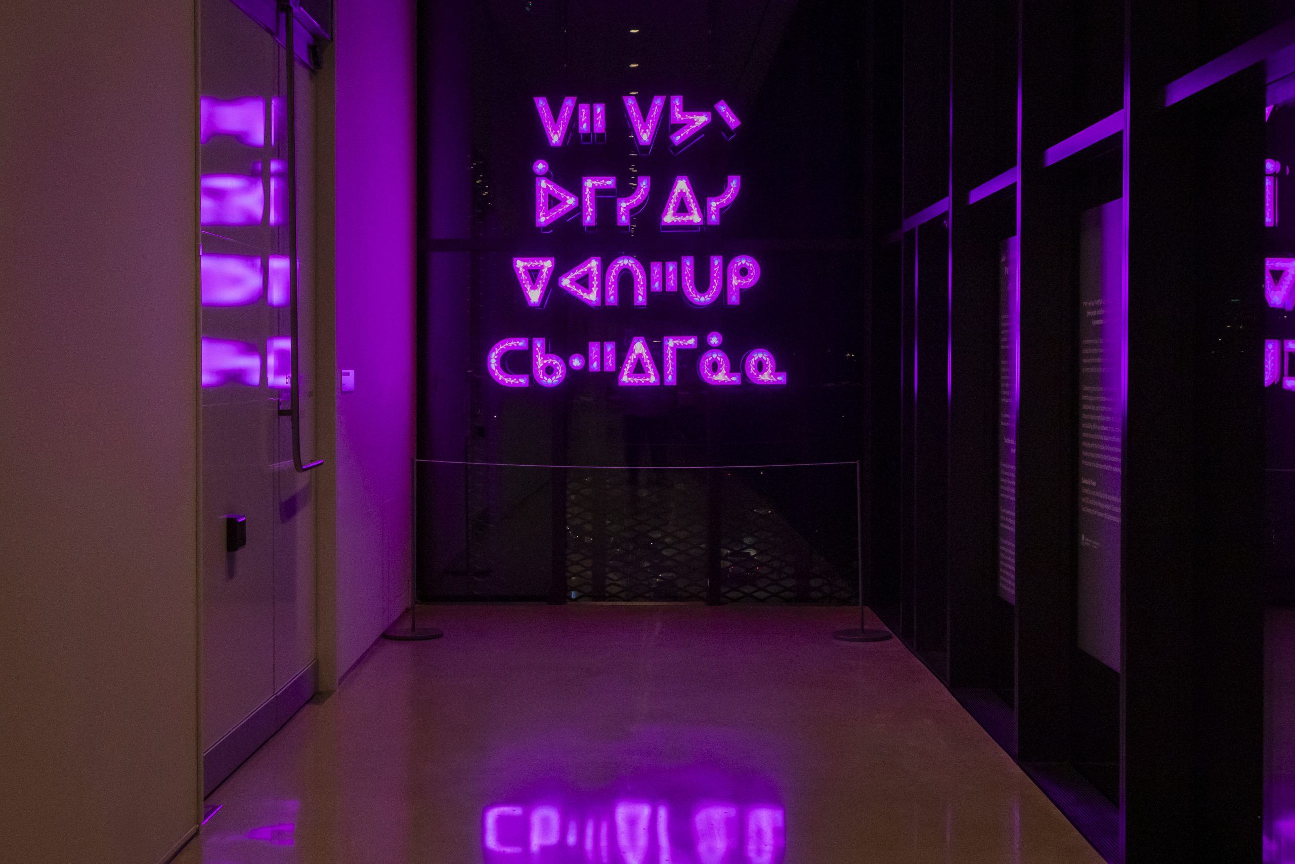 A dimly lit hallway glows with bright magenta light from a neon installation displaying stylized syllabic characters. The glowing text reflects on the polished floor, while the surrounding walls and glass surfaces catch the vivid purple hues.