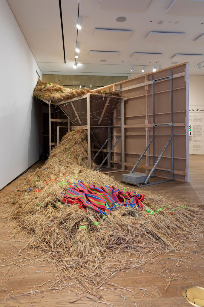 A large-scale sculptural installation made from wheat and exposed wooden framing occupies a contemporary gallery space. The structure resembles an oversized funnel or chute, with straw spilling dramatically from the top and cascading onto the floor in thick piles.Brightly colored ribbons or tape fragments are scattered throughout the straw, and a vivid striped woven textile in red, blue, green, and pink lies draped across the foreground pile. The back of the installation reveals its construction: unfinished plywood panels, timber supports, and metal braces, emphasizing its temporary, architectural quality.