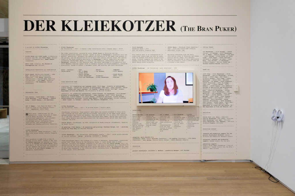 A large exhibition wall text and media installation fills a gallery wall. The headline reads “DER KLEIEKOTZER (The Bran Puker)” in bold black serif lettering across the top. Below it are multiple columns of dense exhibition text, credits, and curatorial information laid out in a structured grid.At the center-right of the wall is a mounted video screen showing a stylized portrait of a woman against a pastel interior background with plants.