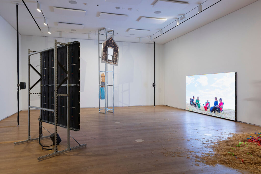 A spacious contemporary gallery installation features multiple sculptural and media elements arranged across a largely open room with wood floors and white walls.On the left stands a tall industrial metal framework supporting a grid of black electronic panels or monitors viewed from the back, with visible cables and structural bracing. Near the center is a narrow vertical steel structure holding a small house-shaped object clad in weathered shingles, with fabric or clothing suspended below it.On the right wall, a large low-mounted video screen displays several people seated outdoors beneath a bright sky, appearing to converse in a field or prairie landscape. Small speakers are mounted around the gallery, suggesting a multi-channel audio installation.