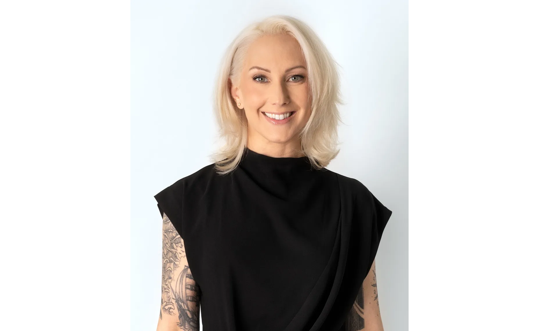 TIna Rivers Ryan headshot A smiling person with short, light-blonde hair wearing a sleeveless black top. They have visible tattoos on both arms and are standing against a plain, light background.