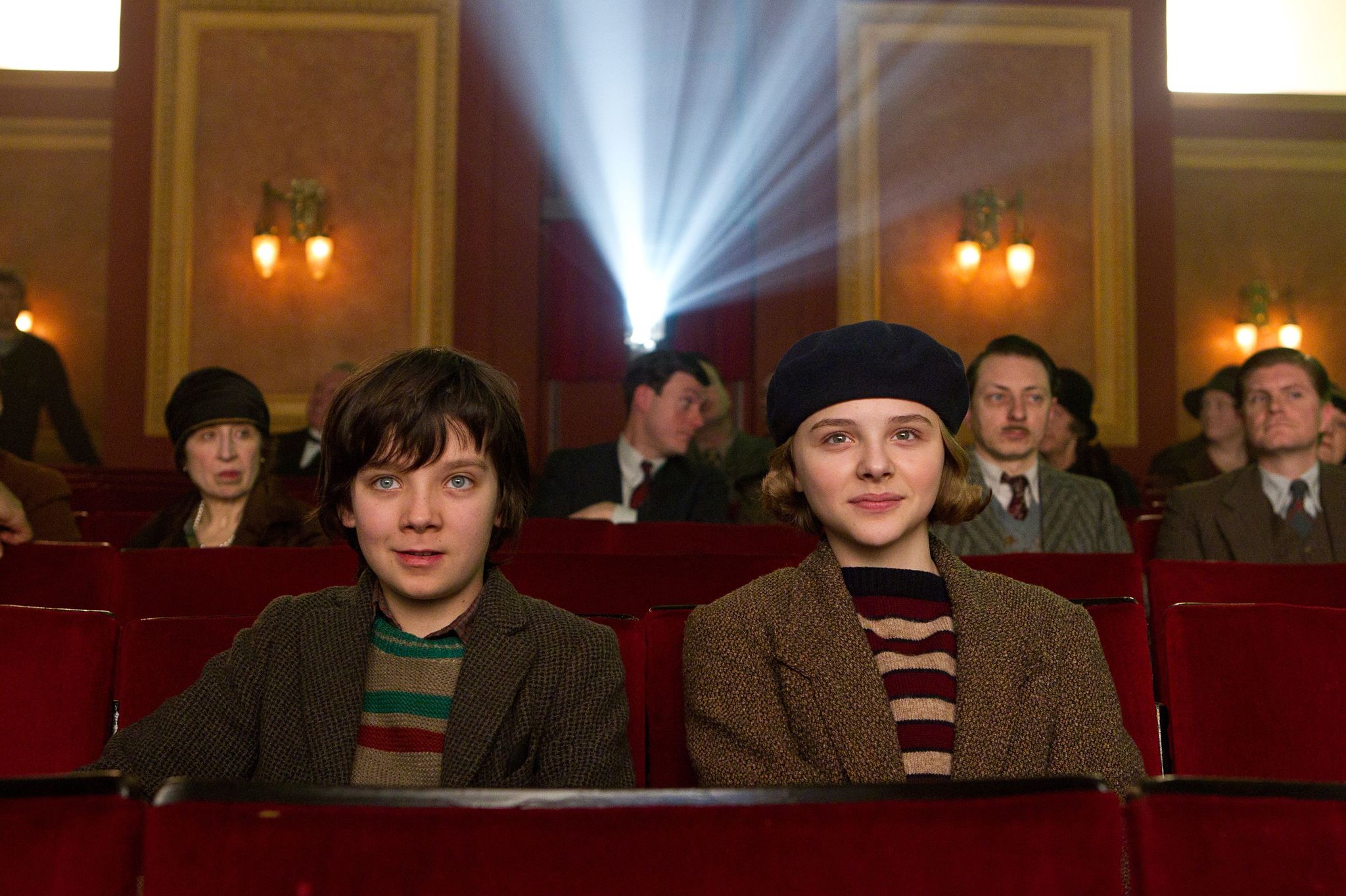 Two children sit side by side in a vintage movie theatre, smiling as they watch a film. A bright beam of light from the projector streams over their heads, illuminating the warm red and gold interior. The boy wears a striped sweater and blazer, while the girl wears a beret and brown coat. Other patrons sit quietly behind them, absorbed in the cinematic experience.