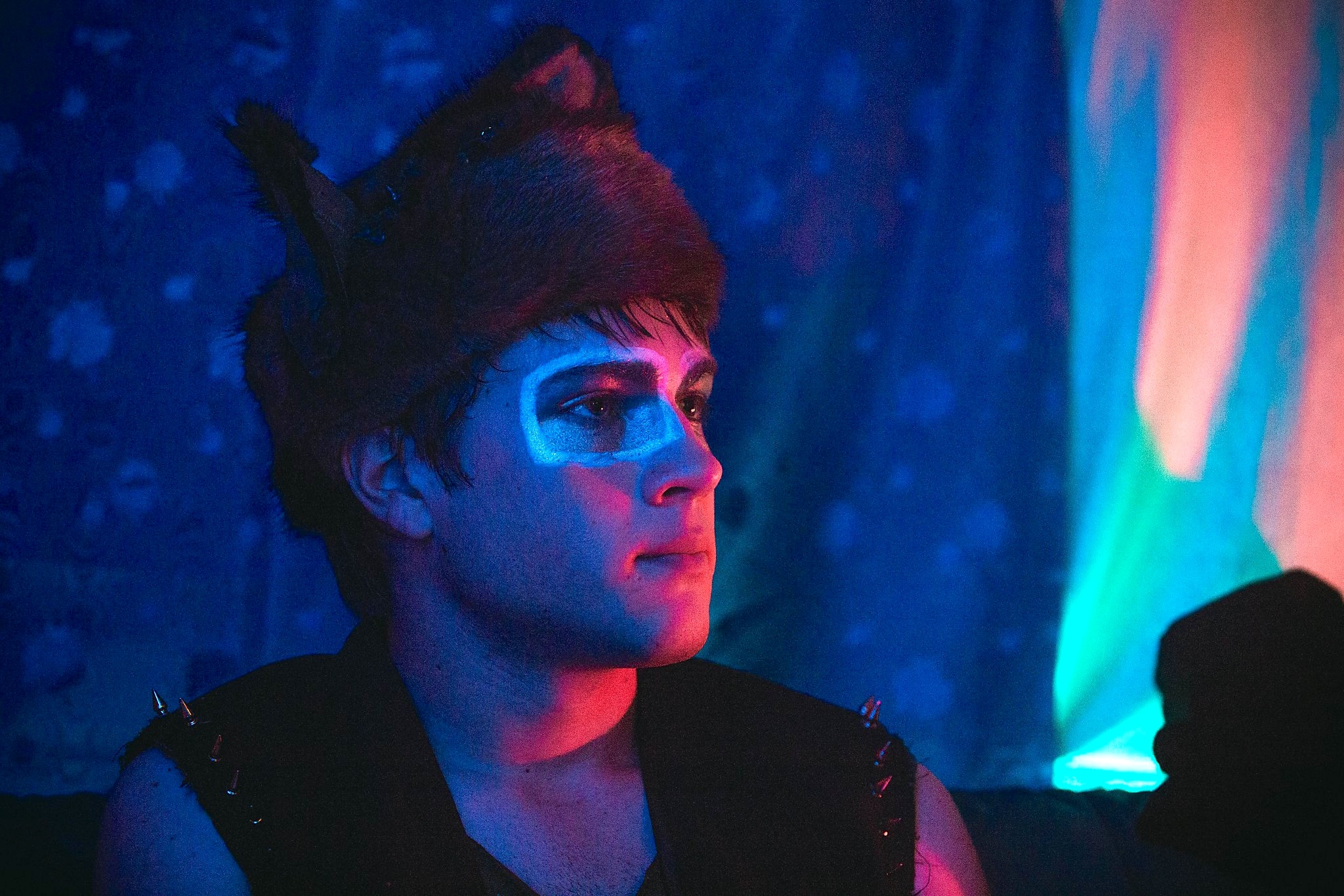 A person wearing a furry hat with pointed ears and bold face paint around one eye looks off to the side in a dimly lit room. Blue, pink, and teal neon lighting washes over their face and the background, creating a moody, nightclub-like atmosphere.