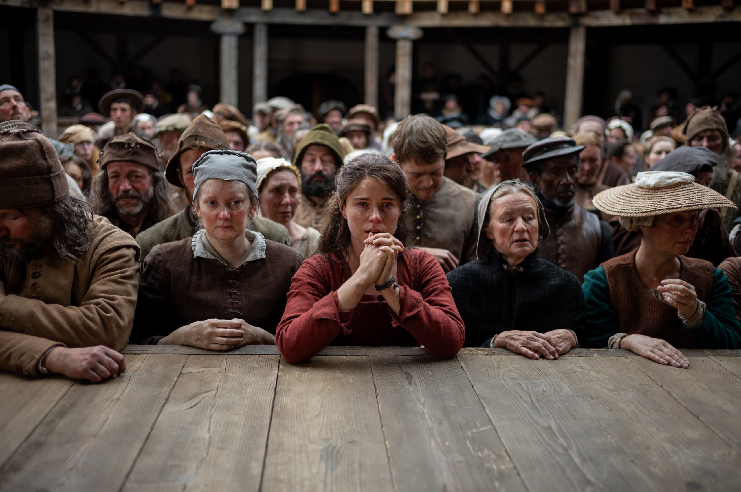 A large crowd of people in simple, historical clothing stands closely packed behind a long wooden table. At the centre, a young woman in a red garment clasps her hands near her face, looking forward with a tense, emotional expression. Others around her—men and women of various ages—look solemn, anxious, or downcast. The setting appears to be an outdoor public gathering in a past era, with wooden structures and muted earth tones.