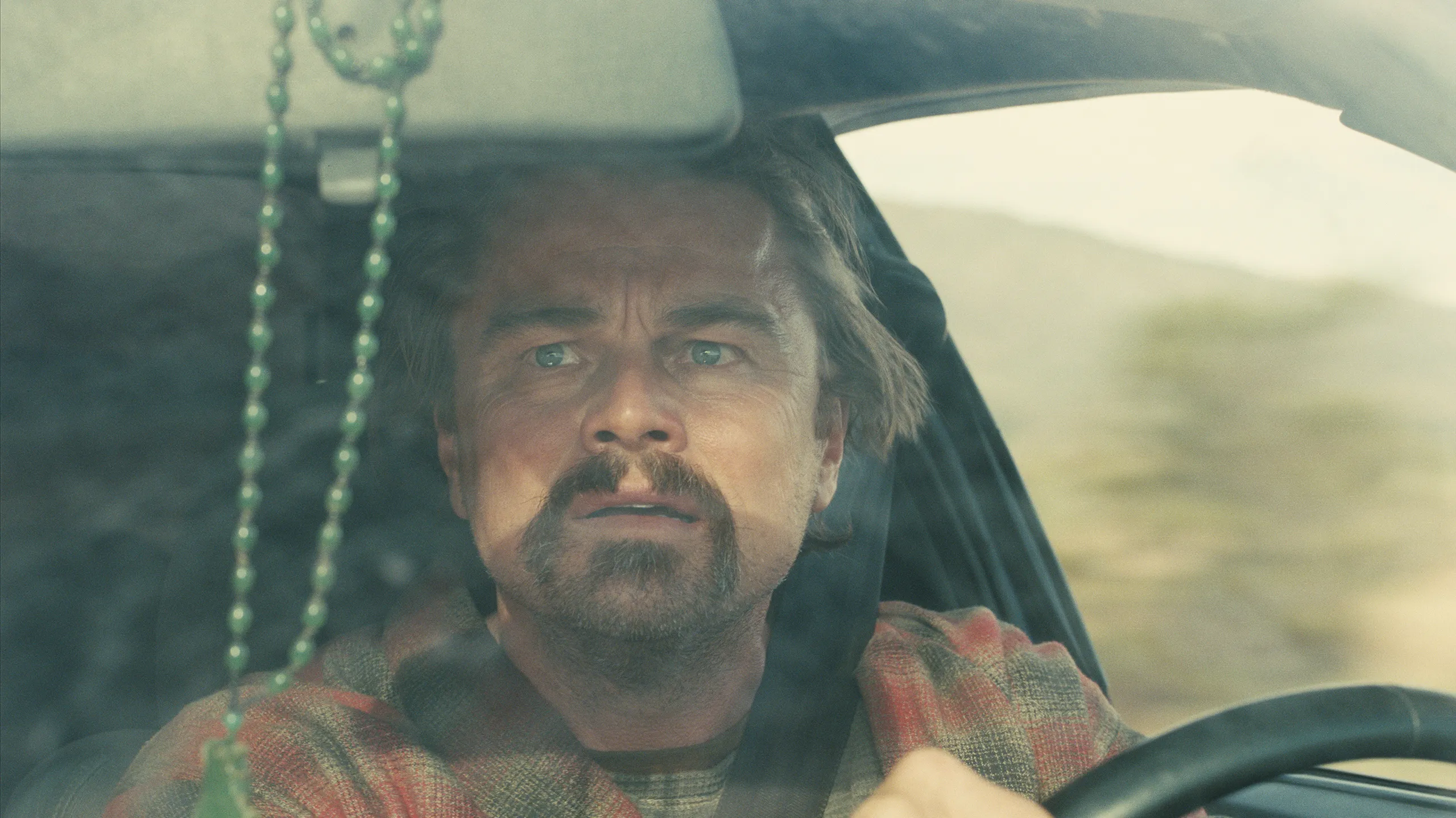 A close-up view through a car windshield shows a middle-aged man driving along an open, grassy landscape. He grips the steering wheel with a tense, focused expression, his eyes fixed straight ahead. A string of green beads hangs from the rearview mirror in the foreground, slightly out of focus, while muted daylight and rolling hills stretch beyond the glass.