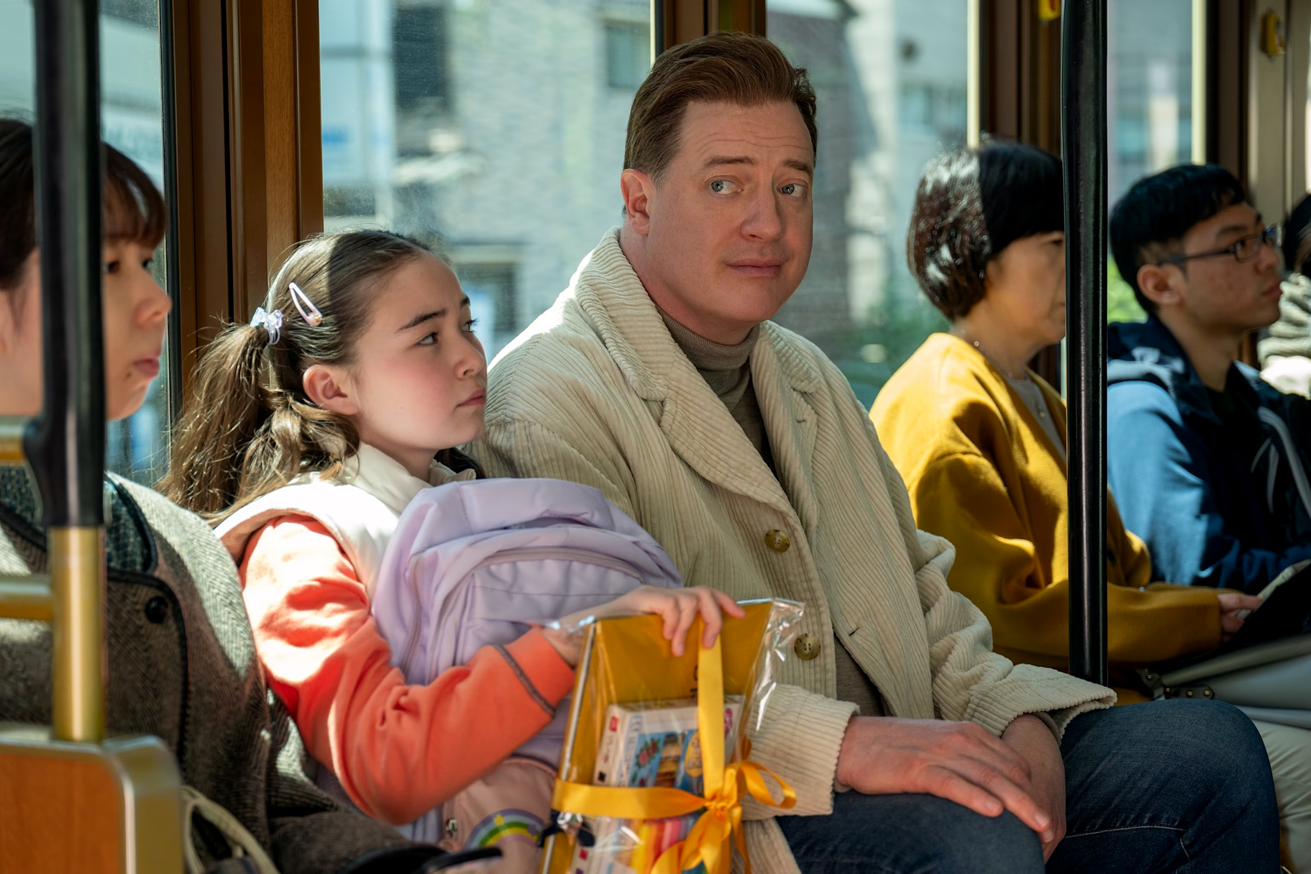A man and a young girl sit side by side on a public transit vehicle, surrounded by other seated passengers. The man wears a light-colored corduroy jacket and looks ahead with a gentle, thoughtful expression. The girl leans close to him, holding a pastel backpack and a clear gift bag tied with a yellow ribbon.