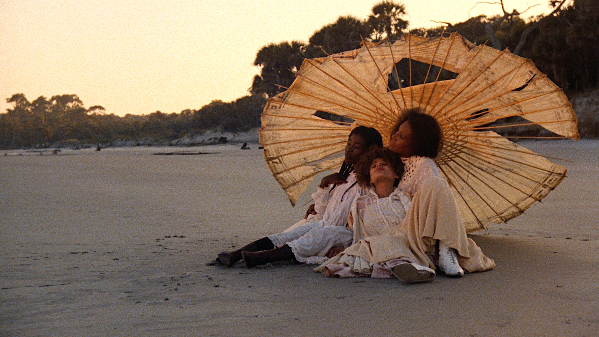 Three people sit close together on a quiet sandy beach at sunset, leaning against one another beneath a large, weathered parasol. They wear light-colored, flowing dresses that blend with the warm tones of the sand and sky. Trees line the shoreline in the distance.