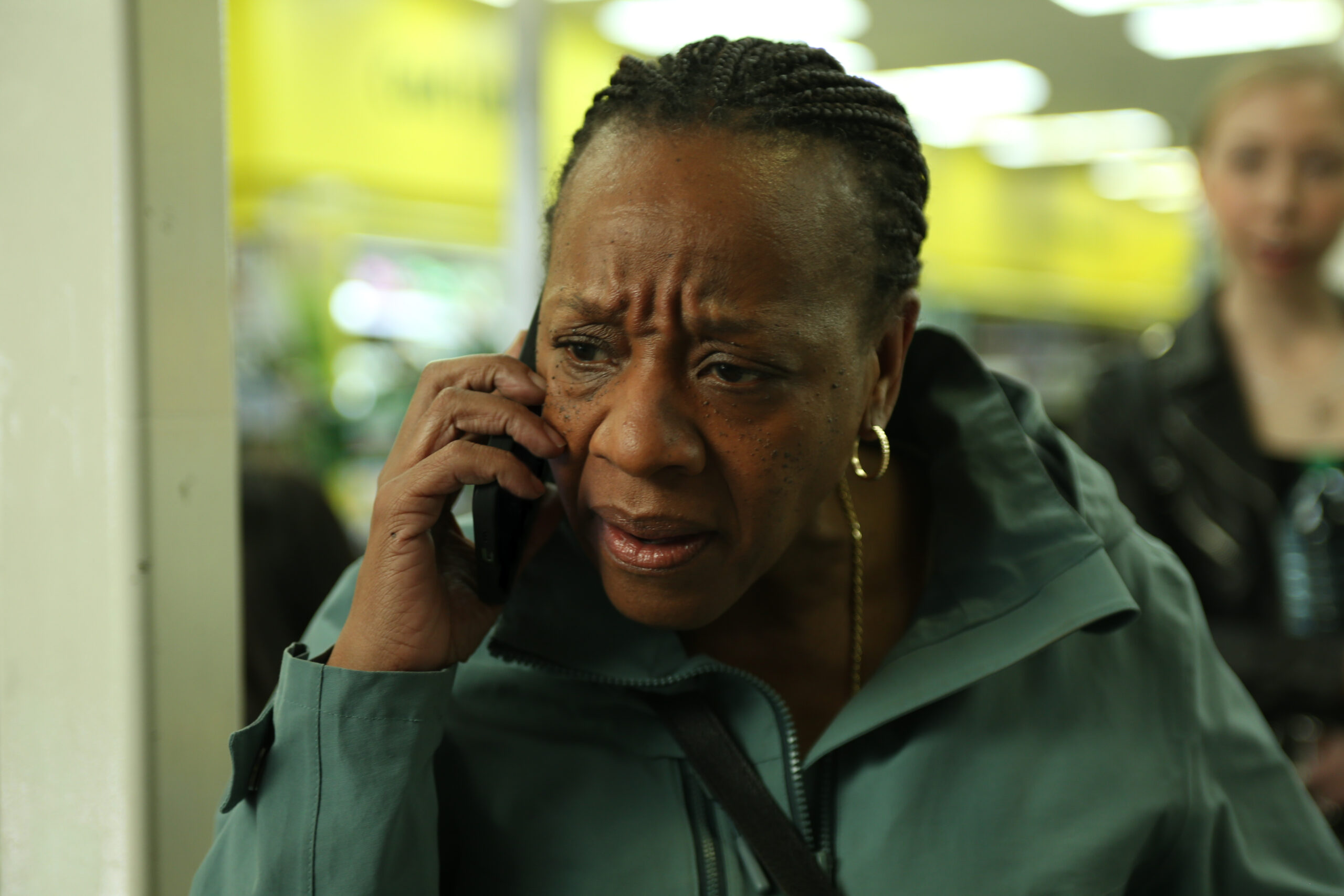 A woman stands indoors in a brightly lit public space, holding a phone to her ear with a worried, tense expression. Her brow is furrowed as she listens closely, wearing a teal jacket and gold hoop earrings. Other people and yellow signage are blurred in the background.