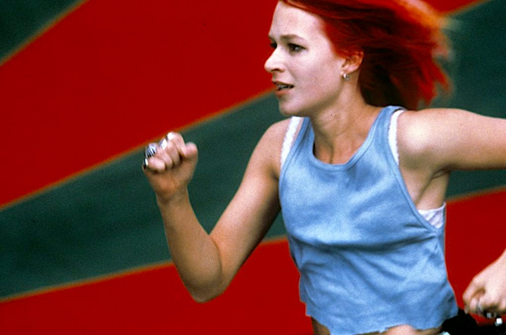 A red-haired woman runs at full speed, her hair streaming behind her. She wears a sleeveless light-blue top and clenches her fist as she moves forward. The background is a blur of bold red and dark green diagonal shapes.