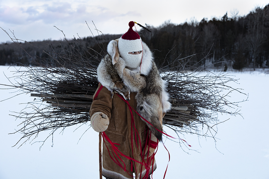 A person stands outdoors in a snowy winter landscape, wearing layered cold-weather clothing including a fur-trimmed coat and a knit face covering. They carry a large bundle of bare branches across their back, secured with red cords, with a wooded area visible in the background.
