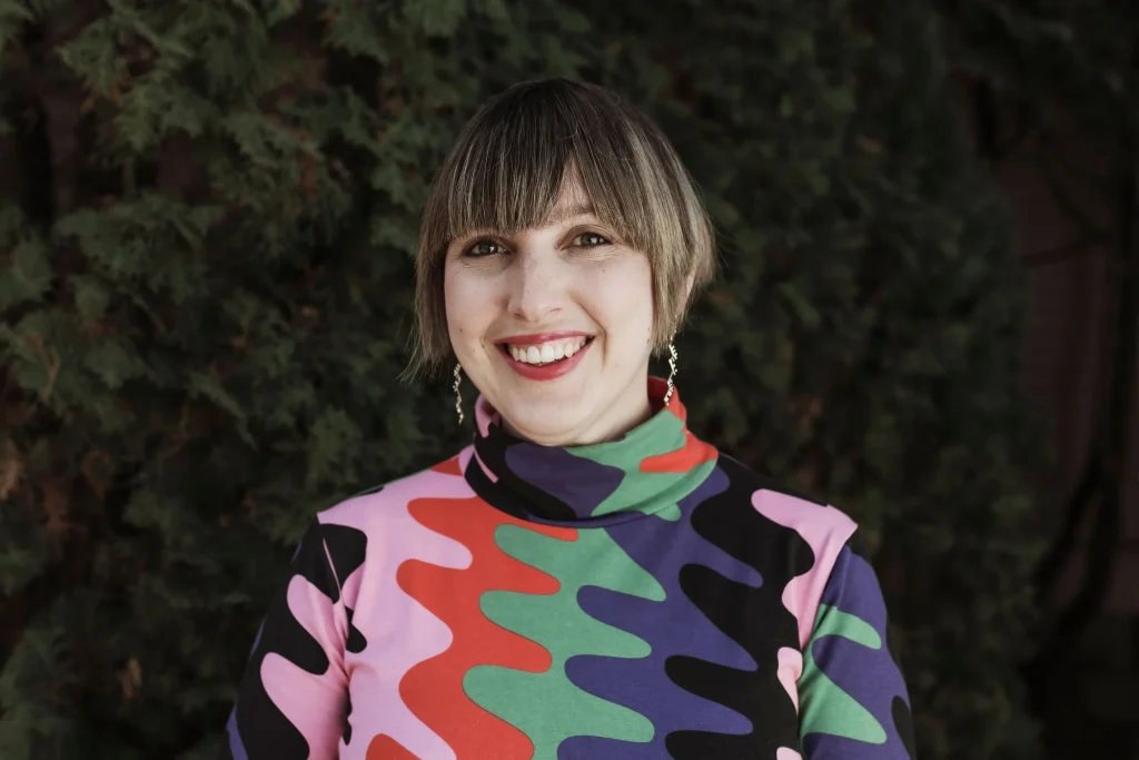 Smiling woman with a short bob haircut and bangs standing outdoors in front of dark green foliage. She wears dangling earrings and a high-neck, long-sleeve top with a bold abstract pattern in pink, red, green, purple, and black.