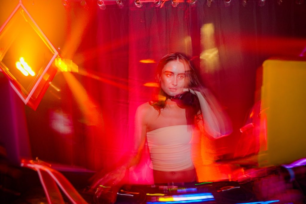 Young DJ standing behind a mixing console in a dimly lit club, bathed in vivid red and orange lights. She wears a strapless top and headphones around her neck, with one hand raised to her ear. Blurred light trails and motion effects create a dynamic, energetic atmosphere, with stage lights and equipment visible around her.