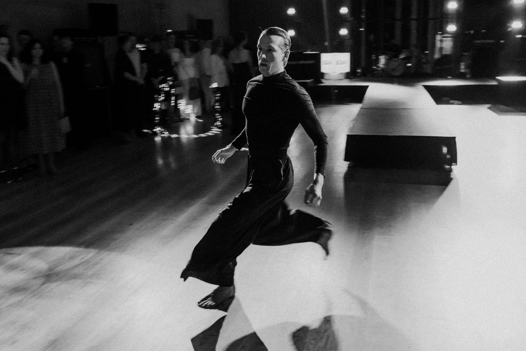 A black-and-white photograph of a dancer moving quickly across a polished floor in a large indoor space. The dancer wears a fitted dark long-sleeve top and wide-legged pants, with hair pulled back. One leg extends forward mid-stride while the other trails behind, creating a sense of motion. In the background, a small audience stands along the edge of the room near a raised platform stage, softly lit by overhead lights. The lighting creates strong highlights and shadows, emphasizing the movement and reflective surface of the floor.
