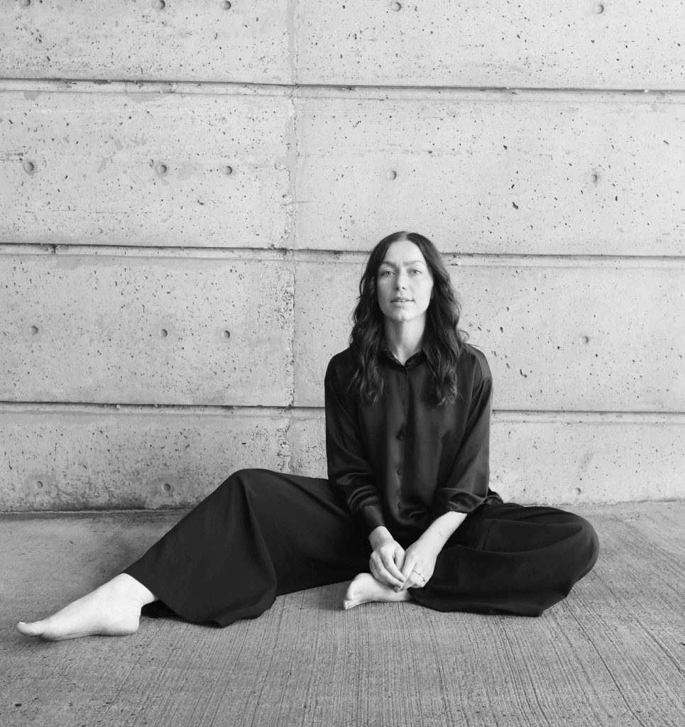 A black-and-white portrait of a person sitting barefoot on a concrete floor against a textured concrete wall. They sit with legs extended wide and slightly bent, wearing loose dark pants and a long-sleeved dark blouse. Their hands rest loosely together in their lap. They look directly at the camera with a calm, neutral expression. The composition is minimal, with strong horizontal lines in the concrete wall creating a clean, modern backdrop.