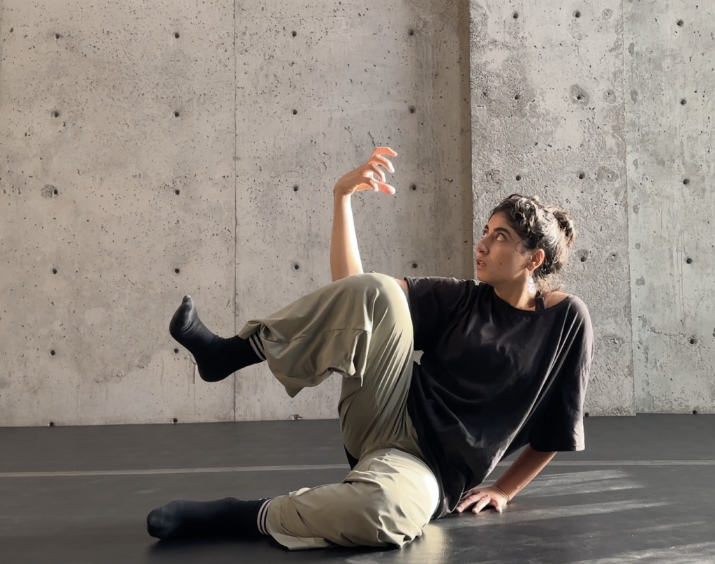 A person sits on a dark studio floor against a raw concrete wall, posed in a contemporary dance position. One leg is bent and lifted, foot flexed in a black sock, while the other leg extends along the floor. They support themselves with one hand behind them and raise the other hand in a curved, expressive gesture, gazing upward toward it. They wear loose beige pants and an oversized black shirt. Soft natural light from the side highlights their face and arm, creating gentle shadows on the textured concrete wall.