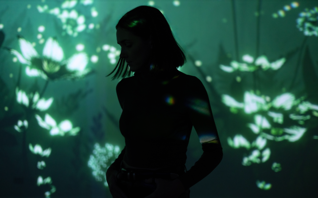 Silhouetted woman in profile standing against a dark backdrop with soft green projected light patterns resembling flowers and glowing shapes cast across the wall and her body, creating a moody, immersive atmosphere.