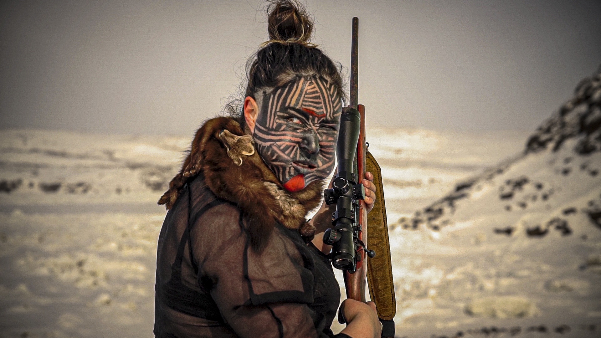 Person standing in a snowy, mountainous landscape holding a scoped rifle over their shoulder. Their face is painted with bold black geometric patterns and a red accent on the chin. They wear a sheer black top and a fur garment draped around their shoulders, looking directly at the camera against the stark, icy backdrop.