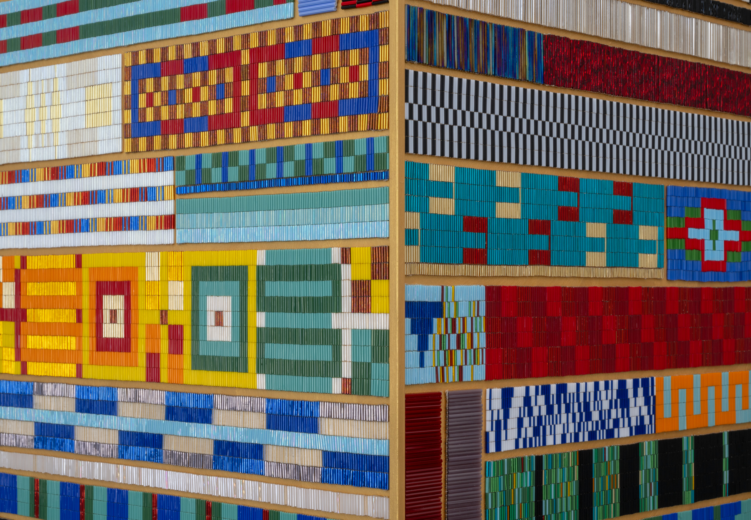 A close-up detail of a large three-dimensional artwork covered in densely packed, vibrantly coloured geometric patterns. The surface is divided into horizontal bands and rectangular sections, each filled with intricate pixel-like arrangements of small tiles or rods in bold colours — red, yellow, blue, teal, green, orange, white, and black. Patterns range from checkerboards and concentric squares to stripes and cross motifs. The corner of the cube-like structure is visible, revealing two faces of the work. Gold-toned framing separates the sections.