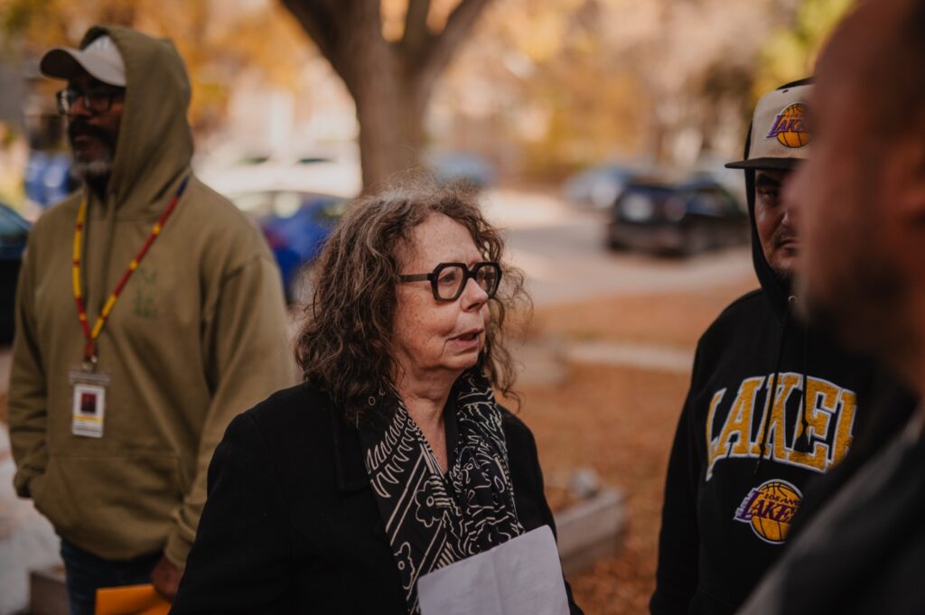 A group of people stand outdoors in autumn, with an older person wearing glasses speaking to others while holding papers.