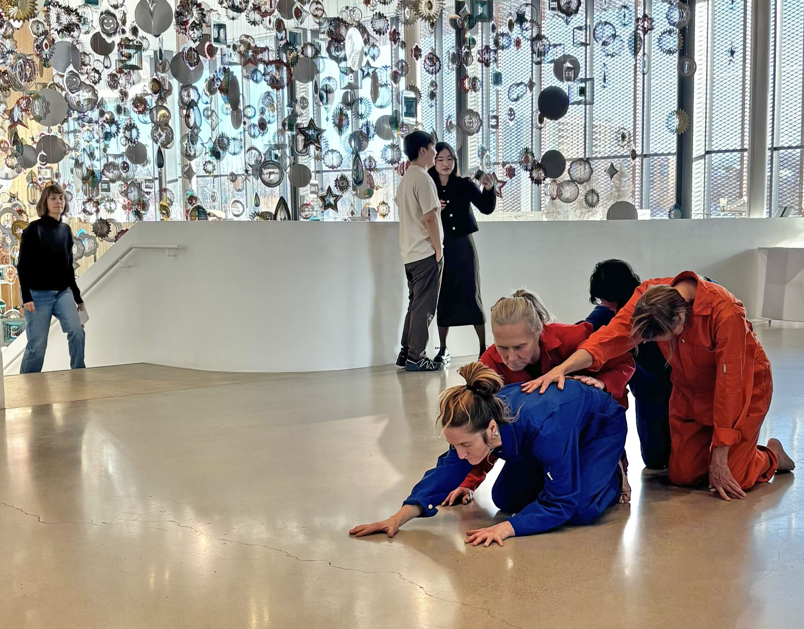 Three performers in bright blue and red outfits kneel and lean forward on a gallery floor while others stand nearby, with a large hanging installation of reflective shapes in the background.