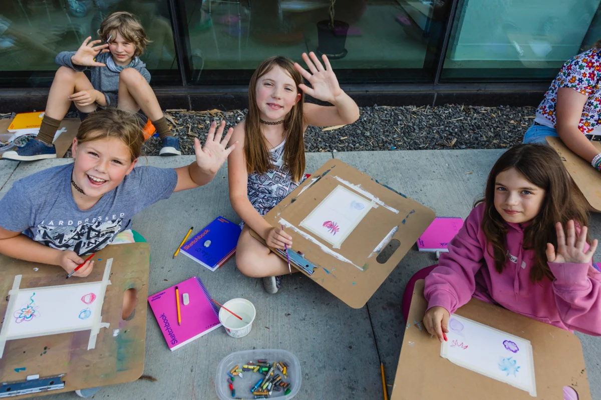 Children sit outdoors on a sidewalk drawing on clipboards, smiling and waving at the camera while surrounded by sketchbooks, crayons, and art supplies.