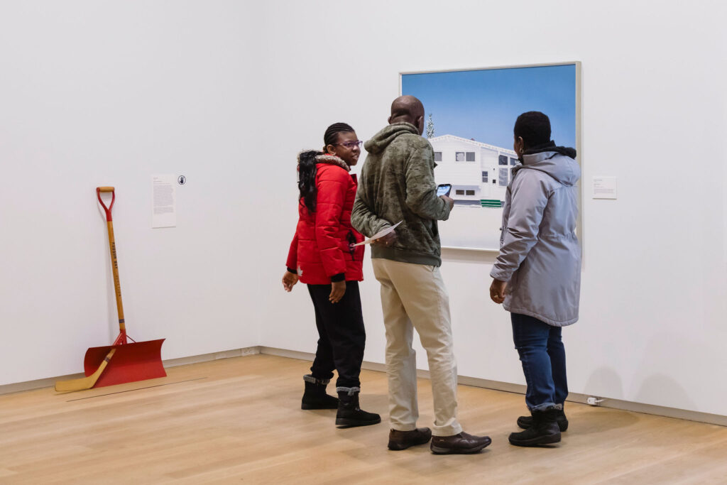 Three visitors gather closely to view a painting of a house, while a red snow shovel rests against the gallery wall nearby.