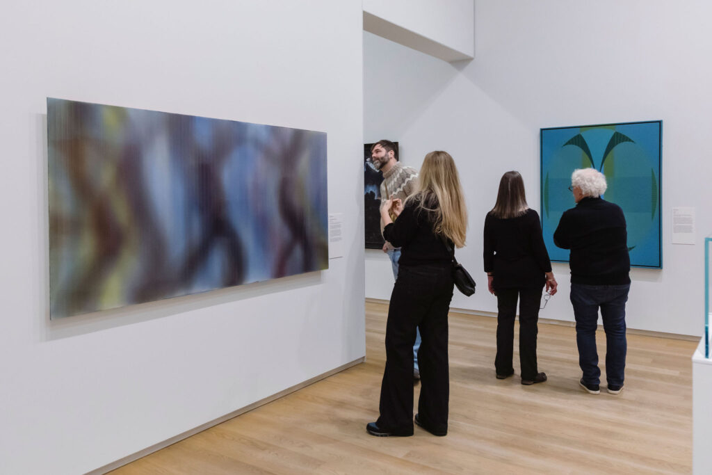 Visitors stand and converse while viewing contemporary paintings in a bright gallery space.