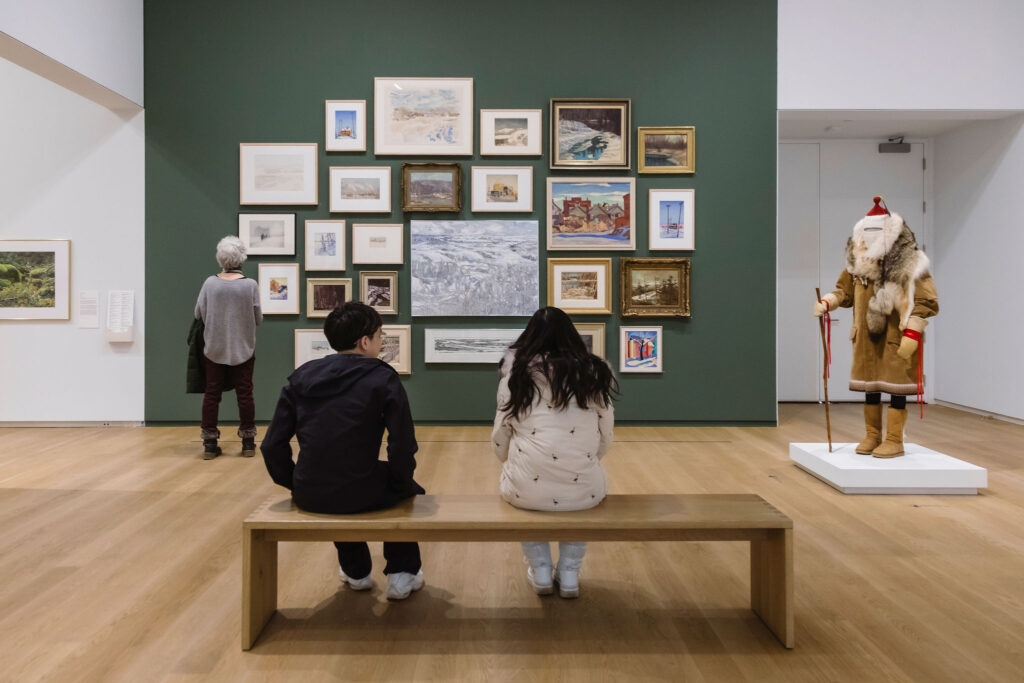 Two visitors sit on a bench viewing a salon-style arrangement of framed artworks on a green gallery wall, while another visitor stands nearby and a sculptural figure is displayed on a pedestal to the side.