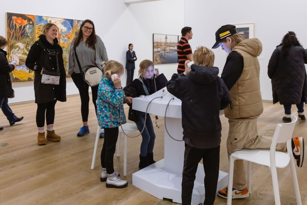 A group of children engage with an interactive audio installation in a gallery, listening through headphones while adults observe nearby.