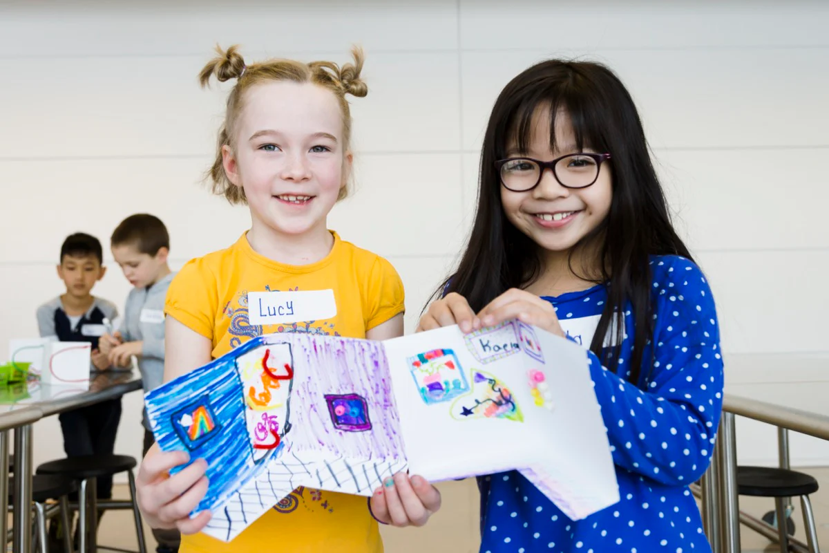 Two children stand indoors smiling at the camera while holding up a colorful handmade booklet of drawings and artwork.