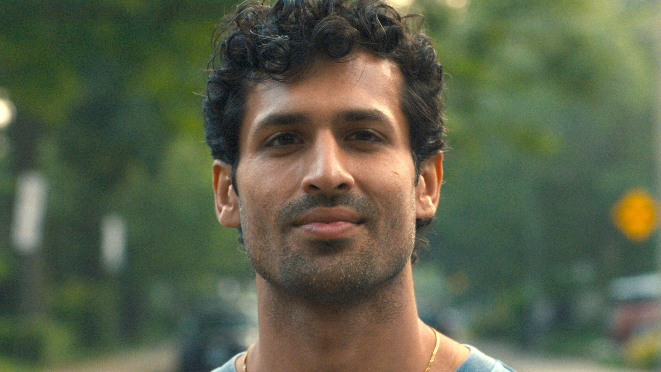 A close-up portrait of a man with short, curly dark hair and light stubble, looking calmly at the camera. He is outdoors, with a softly blurred background of greenery and hints of street elements behind him.