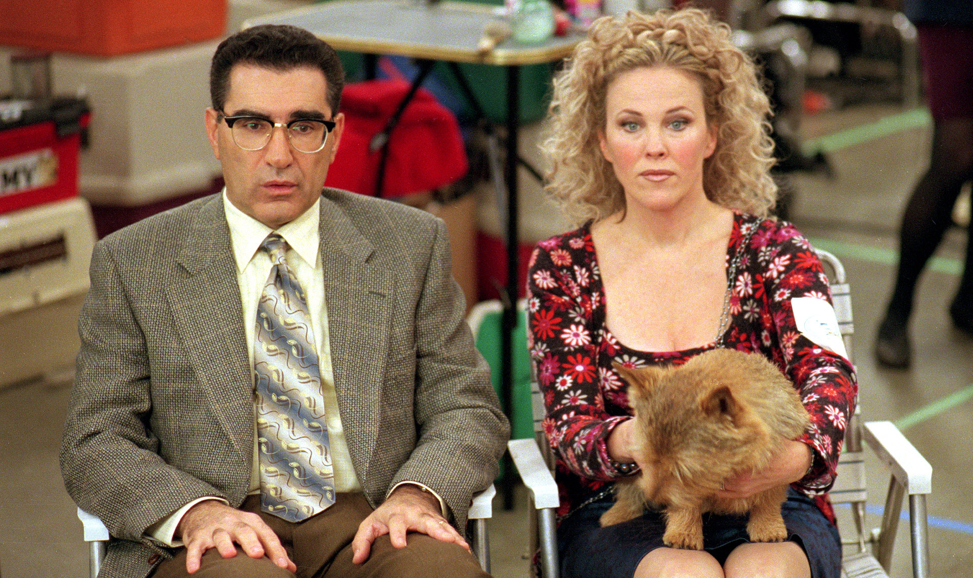 Two people sit side by side at an indoor event. The man in a suit and glasses looks slightly tense, while the woman with curly blonde hair holds a fluffy brown cat out in front of her.