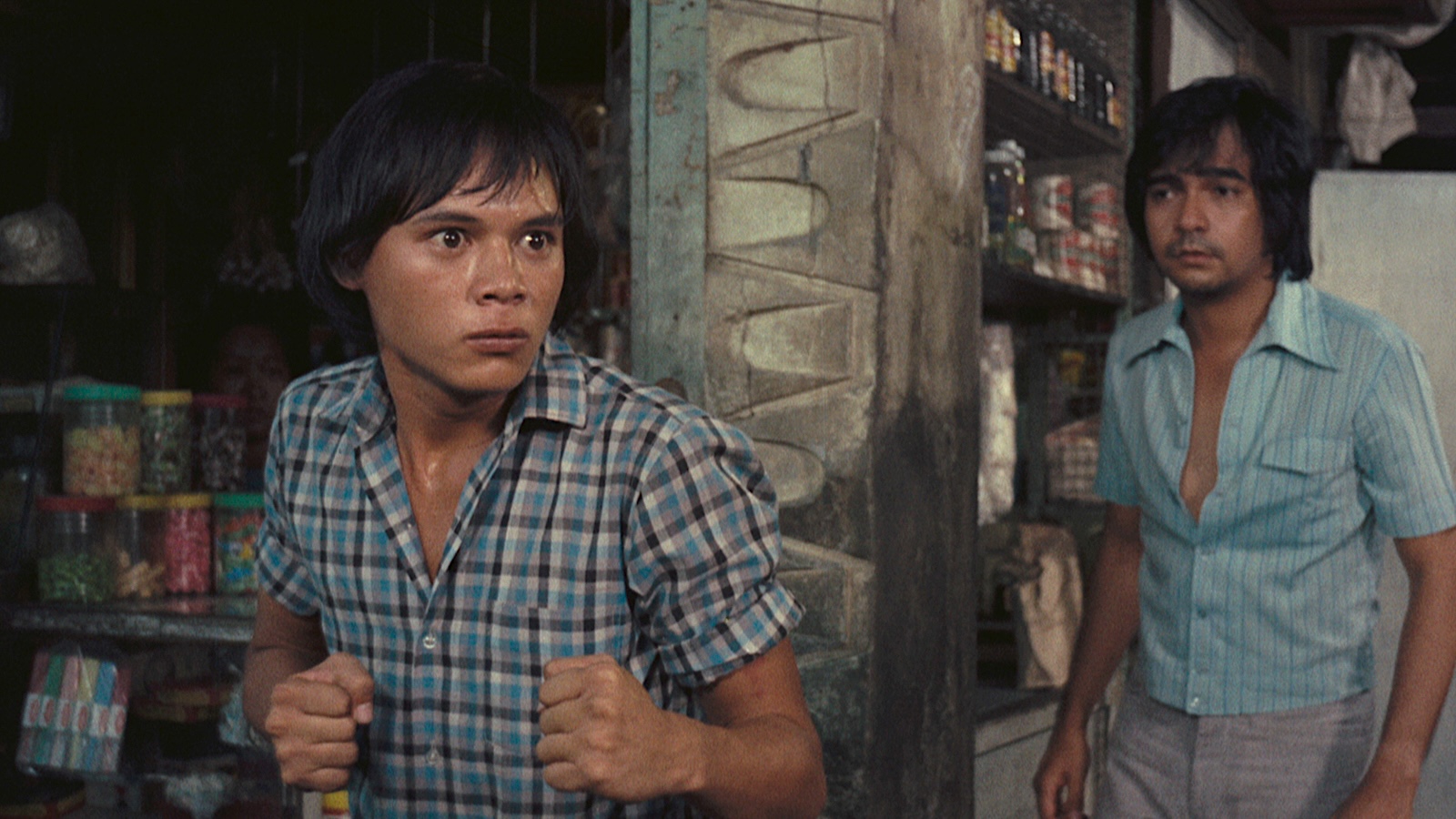 Two young men stand near a weathered doorway of what appears to be a small corner shop or sari-sari store, with shelves of canned goods and jars visible in the background. The man in the foreground wears a blue plaid short-sleeve shirt and holds his fists raised in a defensive stance, his expression tense and alert, while a second man in a light blue shirt looks on warily from behind the doorframe. The warm film grain and styling suggest a movie still from the 1970s or 1980s.